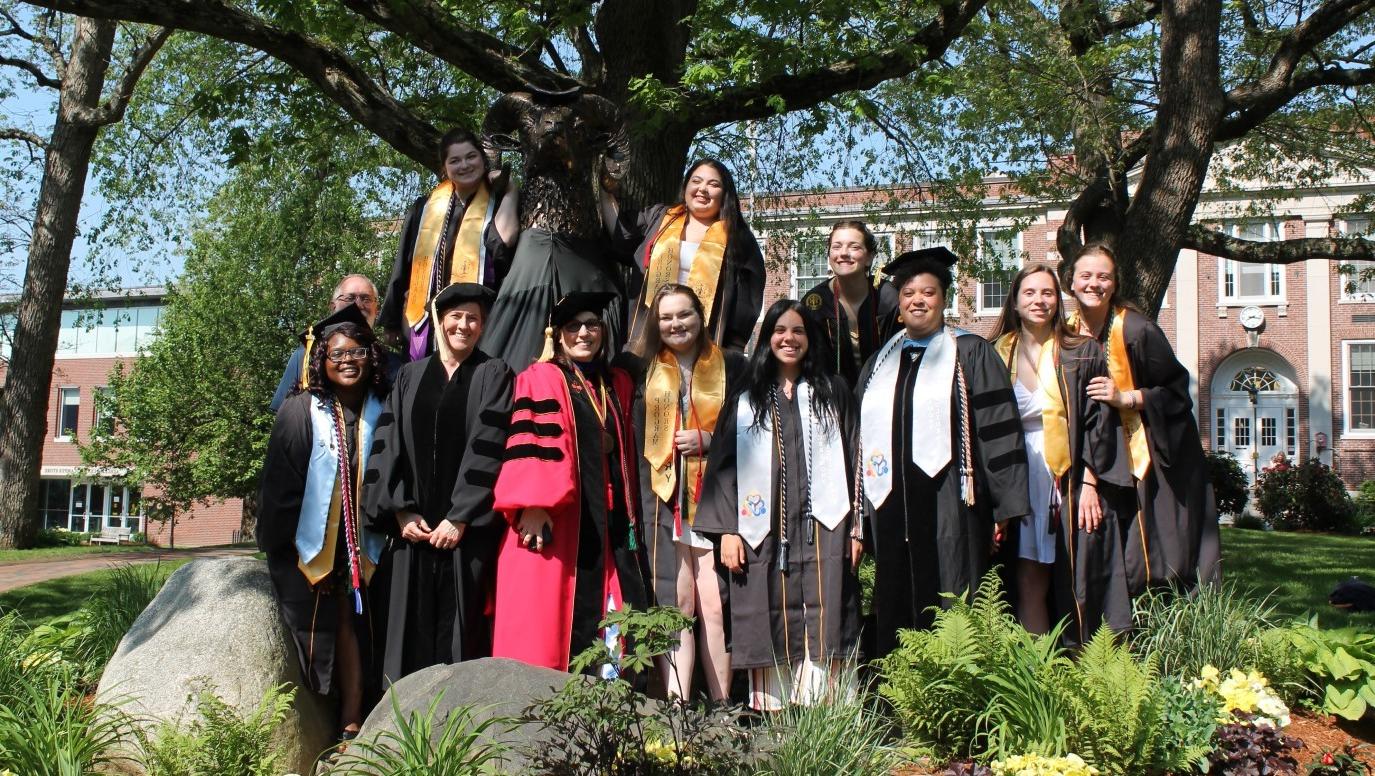 Some 2022 Biology graduates in academic regalia with faculty pose next to the Ram Statue in Crocker Grove